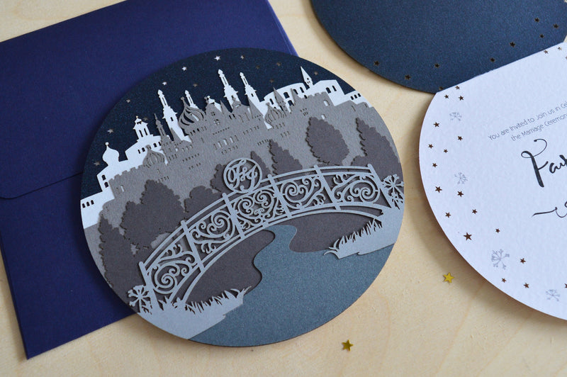 A circular wedding invitation suite displayed on a light wood background with gold star confetti. The set includes a navy blue envelope, a round card featuring a gray and white silhouette of a castle over a bridge, a round dark navy card, and a white oval invitation card with black script text reading 'Paddy & Jasmin'.